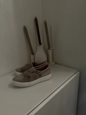 Beige sneakers med rosett - Säljer ett par beige slip-on sneakers med stor rosettdetalj på ovansidan och vit platåsula. Skorna har en mjuk mockaliknande yta och en stilren, modern look. Perfekta för dig som vill ha något både bekvämt och trendigt.