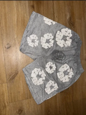 Grå shorts med vit grafisk print, den är blöt när jag tog bilden men den är mycket ljusare - Säljer ett par grå shorts med bred resår i midjan och stora vita grafiska tryck över hela plagget. Shortsen har en loose passform och är tillverkade i mjukt material som känns skönt mot huden. Perfekta för en avslappnad och trendig look.