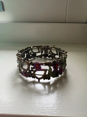 Vintage armband med blommor och blad - Unikt armband i metall med antik finish, dekorerat med lila/rosa blommor samt gröna blad. Armbandet har en öppen, länkad design och detaljerade utsmyckningar som ger en bohemisk känsla. Saknas en röd pärla på en utav blommorna (se bild 4) men det syns knappt💗