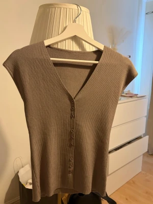 Beige ribbad topp med knappar - Säljer en beige ribbad topp med v-ringning och knappar framtill. Toppen är ärmlös och har en slim passform som sitter snyggt på kroppen. Perfekt att styla med jeans eller kjol för en trendig look.