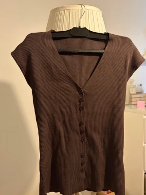 Brun ribbad v-ringad topp - Säljer en brun ribbad topp med v-ringning och knappar framtill. Toppen har korta ärmar och är tillverkad i ett mjukt, stretchigt material som sitter snyggt på kroppen. Perfekt att styla med jeans eller kjol för en trendig look.