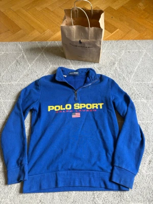 Blå polo Ralph lauren tröja (polo sport)  - Blå långärmad tröja från Polo Sport Ralph Lauren med halv dragkedja och stor gul logga framtill. Tröjan har ribbade muddar och mjukt material på insidan. Perfekt för dig som gillar sportig och klassisk stil. Den har ett litet håll men inget som märks (syns på sista bild) Påse ingår såklart. 