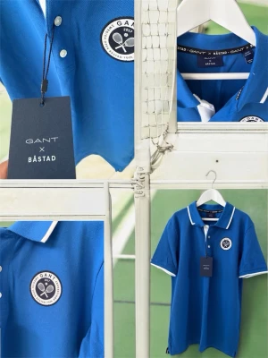 Blå GANT x Båstad pikétröja - Blå pikétröja från GANT x Båstad med vit krage och vita detaljer på ärmsluten. Tröjan har en broderad patch med tennismotiv på bröstet och klassisk pikéknäppning. Perfekt för en sportig och stilren vibe. Nypris 599