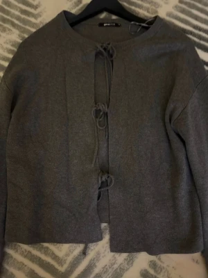 Grå kofta med knytning från Gina Tricot - Trendig grå kofta från Gina Tricot med tre knytband framtill. Koftan har rund halsringning och är gjord i mjukt stickat material. Perfekt att styla över ett linne eller t-shirt för en chill look. Aldrig använd. Xs men passar S
