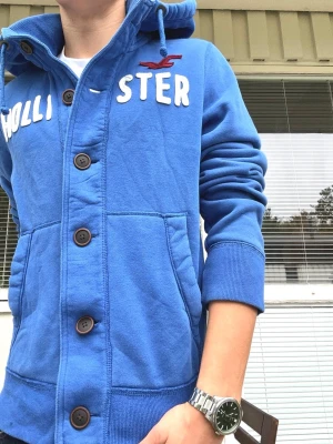 Sällsynt Hollister Vintage Hoodie Cardigan M eller L - En extremt härlig hollister hoodie i sällsynt modell🤩 Blå färg och snygga knappar längs med framsidan🙌 Hoodien är i vintage och archive stil, perfekt nu till hösten och vintern🍁❄️ Kvaliteten är tydligt framstående och det tjocka tyget gör att plagget sitter riktigt fint🤩 Inga defekter, nyskick i princip🤝 Extremt snygga fabriksgjorda slitningar på hollister - texten🙌 Stl L, men passar nog många med M också👍 Längd från axel: 66cm. Först till kvarn🤩