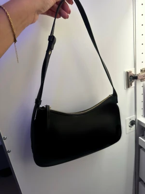 Svart axelremsväska i skinnimitation - Stilren svart handväska med justerbar axelrem och guldfärgad dragkedja. Mjukt rundad form och är tillverkad i skinnimitation. Perfekt för dig som gillar minimalistisk och trendig design.