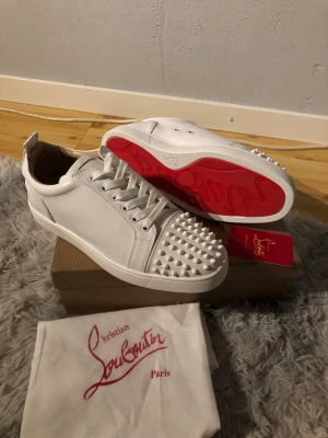 Vita Christian Louboutin sneakers - Säljer ett par vita sneakers från Christian Louboutin med ikoniska röda sulor och nitar på tån. Skorna är låga och har vita skosnören samt detaljer i läder. Perfekta för dig som vill sticka ut med en lyxig och edgy stil.