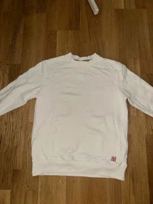 Tröja Fram - En enkel och stilren vit sweatshirt med rund halsringning och långa ärmar. Tröjan har ribbade muddar vid ärmslut och nederkant samt en liten röd detalj nertill på framsidan. Perfekt för en clean och avslappnad look. Det står L men passar både M