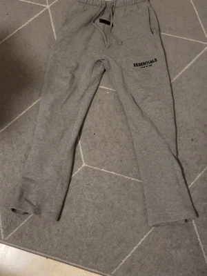 Grå Essentials Fear of God mjukisbyxor - Säljer ett par grå mjukisbyxor från Essentials Fear of God. Byxorna har dragsko i midjan, raka ben och fickor på sidorna. Tillverkade i mjukt sweatshirtmaterial och har logga tryckt på vänster lår. Perfekta för en avslappnad stil.