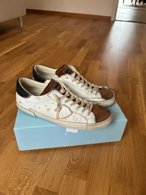 Philippe Model vita och bruna sneakers - Snygga sneakers från Philippe Model i vitt läder med bruna mockadetaljer på tån och plösen. Skorna har svarta detaljer vid hälen och en ikonisk sköld på sidan. Klassisk låg modell med vita snören och slitstark sula.