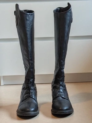 RIDSTÖVLAR CRW , JUNIOR CLIFTON  - Snygga svarta ridstövlar i läder med klassisk snörning framtill och dekorativa sömmar. De har en robust sula med bra grepp och en knappdetalj upptill. Perfekta för dig som vill ha en stilren och tidlös look. Två små mindre defekter finns på ridstövlarna. Dem två små  defekterna visas på 2 bilder  ( 2 sista bilderna)  Dem två små defekterna fanns när jag köpte ridstövlarna på Hööks och jag fick 50 kr rabatt av Hööks för dem 2 små defekterna som visas på dem 2 sista bilderna.