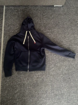 Mörkblå hoodie från Polo Ralph Lauren - Snygg mörkblå hoodie från Polo Ralph Lauren med dragkedja, vita snören i huvan och röd logga på bröstet. Mjuk fleece på insidan och två stora fickor framtill. Perfekt för en chill och stilren look.