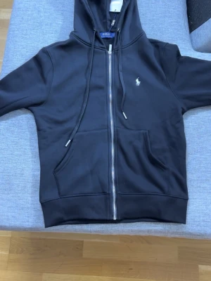 Svart hoodie från Polo Ralph Lauren - Svart hoodie med dragkedja från Polo Ralph Lauren,storlek M.Klassisk design med huva och dragsnören, två fickor framtill och den ikoniska vita logon broderad på bröstet. Tillverkad i mjukt material som känns skönt mot huden. Perfekt för en avslappnad och stilren look.