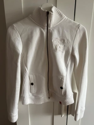 Vit zip-hoodie med broderi - Snygg vit hoodie från edc med dragkedja och ribbad hög krage. Broderad detalj på bröstet och två fickor framtill med knappar. 