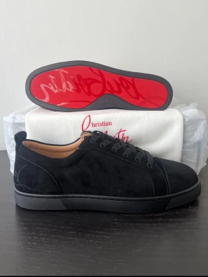 Svarta sneakers Christian Louboutin - Snygga svarta sneakers i mocka från Christian Louboutin med den ikoniska röda sulan och broderad logga på hälen. Skorna har svarta skosnören, låg profil och en stilren design som passar till det mesta. Insidan är fodrad i beige läder för extra komfort. Allt OG medföljer förutom OG box