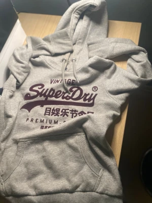 Grå Superdry hoodie med tryck  - Snygg grå hoodie från Superdry med stor vinröd logga och text på bröstet. Klassisk känguruficka framtill och justerbar huva med dragsko. Mjuk bomullsblandning som känns skön mot huden. Perfekt för en chill och avslappnad stil. Skriv vid frågor eller funderingar, är villig med byte (speciellt skor) 