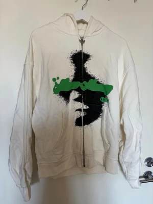 Vit hoodie med grön och svart print - Säljer en vit hoodie med dragkedja och huva. More money more love. 