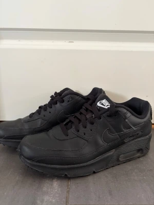 Nike Air Max 90 helsvarta sneakers - Nike Air Max 90 sneakers i helsvart färg med klassisk siluett. Skorna har ovandel i läder och textil, synlig Air-enhet i sulan och svart sula. Ikonisk Nike-logga på plösen och sidan. Perfekta för dig som gillar stilrena och tidlösa sneakers.