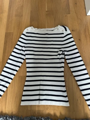 Vit och marinblå randig stickad tröja - Säljer en ribbstickad tröja med vita och marinblå horisontella ränder. Tröjan har långa ärmar, rund halsringning och dekorativa knappar på axlarna. Materialet är mjukt och stretchigt, perfekt för en avslappnad men stilren look.
