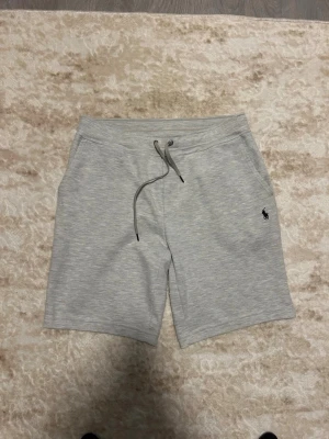 Ljusgrå shorts med snörning - Snygga ljusgrå shorts med snörning i midjan och sidofickor. Klassisk design med diskret logga på vänster ben. Perfekta för avslappnade dagar och enkel att matcha med olika plagg.