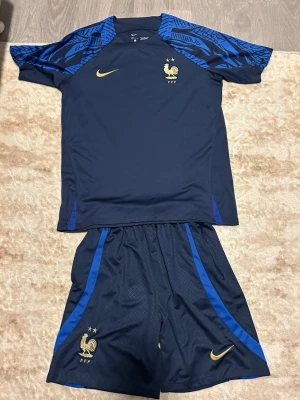 Frankrike fotbollströja och shorts Nike - Mörkblått fotbollskit från Nike med Frankrikes landslagslogga och guldiga detaljer. Tröjan har blå mönstrade axelpartier och korta ärmar, shortsen har blå ränder på sidorna. Perfekt för träning eller match.