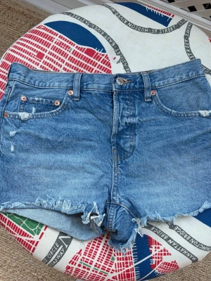 Blå denimshorts med fransig kant - Säljer ett par klassiska blå jeansshorts i bomull med fransig, rå kant och slitna detaljer. Shortsen har fem fickor, bälteshällor och knappgylf. Perfekta för sommaren och ger en avslappnad vibe.