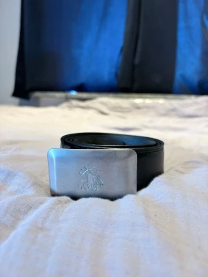 Svart läderbälte Tiger of Sweden - Snyggt svart bälte från Tiger of Sweden i äkta läder. Bältet har ett stilrent, rektangulärt spänne i silverfärgad metall med Tiger of Sweden-loggan ingraverad. Perfekt accessoar för att lyfta din outfit.