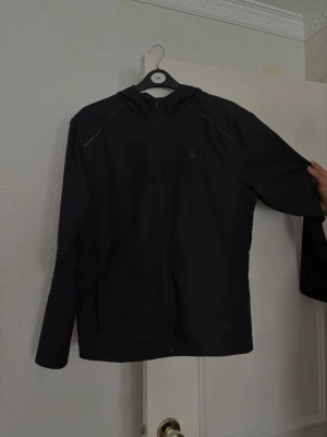 Svart vindjacka med huva - Säljer en svart vindjacka med huva och dragkedja framtill. Jackan har långa ärmar och en enkel, stilren design som passar perfekt till vardags. Tillverkad i lätt och smidigt material, perfekt för blåsiga dagar.