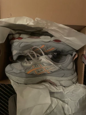 Gråa Asics sneakers med orange detaljer - Snygga sneakers från Asics i ljusgrått med mesh och syntetpaneler. Skorna har orange och silverfärgade detaljer på sidan samt vita skosnören. Sulan är chunky med röda inslag och ger en sportig vibe. Perfekta för dig som gillar retrostil och komfort.