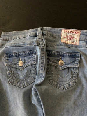 Ljusblå bootcut jeans True Religion - Säljer ett par ljusblå bootcut jeans från True Religion med ikoniska bakfickor och silverfärgade knappar. 