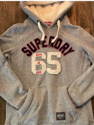 Grå Superdry hoodie med teddyfoder - Mysig grå hoodie från Superdry med stor broderad logga och siffran 65 på bröstet. Utrustad med huva som har vitt teddyfoder och vita dragsnören. Känguruficka framtill och ribbade muddar. Perfekt för chill dagar eller när du vill ha det extra bekvämt.