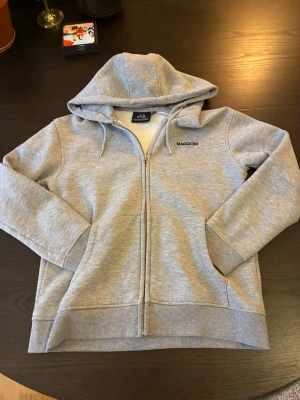 Grå zip hoodie från Maggiore - Snygg grå hoodie från Maggiore med dragkedja framtill och broderad logga på bröstet. Mjuk insida i fleece, två fickor och justerbar huva med snören. Perfekt för chill dagar eller när du vill ha något bekvämt och stilrent.