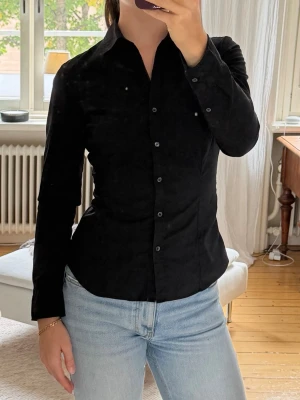 Svart manchester skjorta - Säljer en svart skjorta i mjuk manchester med klassisk krage och knappar framtill. Skjortan har lång ärm och en figurnära passform som ger en snygg siluett. 