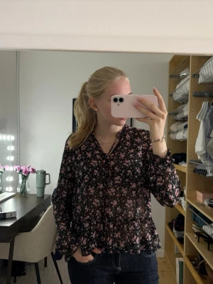 Svart blommig blus med volang - Säljer en svart blus med rosa och vita små blommor. Blusen har långa ärmar, volangkant nedtill och vid ärmslut samt knytning vid halsen. Materialet är tunt och lätt transparent, vilket ger en luftig känsla.