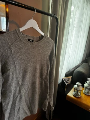 Grå stickad tröja från Uniqlo - En stilren grå stickad tröja från Uniqlo med rund halsringning och ribbade muddar vid ärmslut och nederkant. Tröjan har en klassisk passform och är tillverkad i mjukt material som passar perfekt till lager på lager.