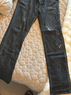 Svarta distressed jeans med DSQ2-tryck - Säljer ett par svarta jeans med slitningar och distressed detaljer. På baksidan finns ett stort orange DSQ2-tryck på ena bakfickan. Jeansen har raka ben och klassisk femficksmodell. Perfekta för en edgy och trendig look. köpt på johnells slutsålda överallt typ! väldigt snygga raka jeans! storlrk 54 i midjan men passar folk sommhar 34/32 med bälte!  nypris runt 6000 ca !