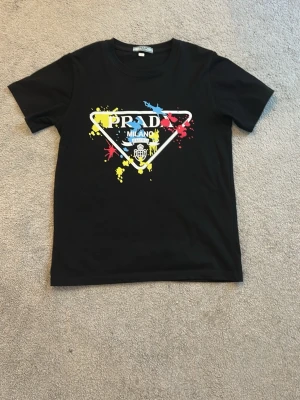 Svart Prada t-shirt med färgstänk och tryck - Prada tröja, svart, på 3e bilden ser man ett vääldigt litet streck syns när ja tar bilden så men i verkligheten syns de verkligen inte. Men därav reducerat pris.