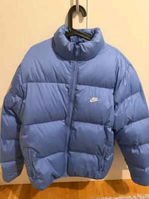 Blå pufferjacka från Nike - Snygg blå pufferjacka från Nike med hög krage och dragkedja framtill. Jackan har en enkel design med diskret Nike-logga på bröstet och två sidofickor med dragkedja. Perfekt för kalla vinterdagar och riktigt skön att bära. Den är knappt använd, användes bara under en vinter, kan ha lite smutsig krage och lite vid handledarna, men kan säkert gå bort i tvätten, inget heller man ser!💙