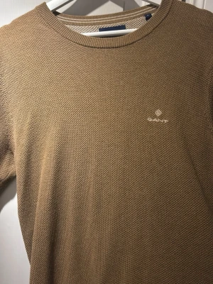 Beige stickad tröja från GANT - Snygg beige stickad tröja från GANT med rund halsringning och diskret logga broderad på bröstet. Tröjan har en klassisk passform och är tillverkad i ett mjukt, strukturerat material som passar perfekt till alla tillfällen.