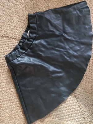 Svart kort kjol i skinnimitation H&M - Snygg svart kort kjol från H&M i skinnimitation. Kjolen har en elastisk midja med knapp och är perfekt för att skapa en edgy look. Passar till många olika stilar och är enkel att matcha med både t-shirt och stickad tröja.