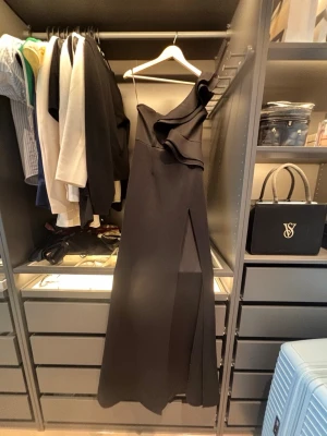 Svart lång bal och festklänning - Elegant svart långklänning med asymmetrisk one shoulder-design och volangdetalj vid axeln. Klänningen har endast användts en gång och är i väldigt bra skick. 