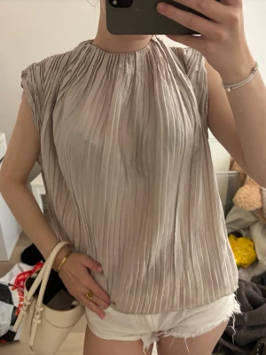 Beige plisserad blus med rund hals - Säljer en beige plisserad blus med rund hals och ärmlös design. Toppen har ett luftigt fall och fina veck som ger en elegant look. Perfekt till sommaren och lätt att matcha med olika stilar.