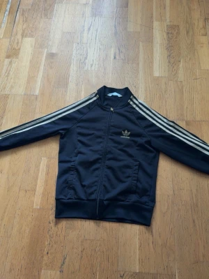 Svart Adidas bomberjacka med guld ränder - Snygg svart bomberjacka från Adidas med klassiska vita ränder längs ärmarna och dragkedja framtill. Jackan har Adidas logga på bröstet och ett stort broderat märke på ryggen. Perfekt för en sportig och avslappnad stil.