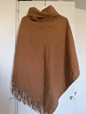 Brun poncho med fransar - Säljer en snygg brun poncho med polokrage och långa fransar längs nederkanten. Ponchon är mjuk och har en lös passform som passar perfekt över en tröja. Materialet känns som en varm och skön ullblandning. Perfekt för lager på lager-stil.