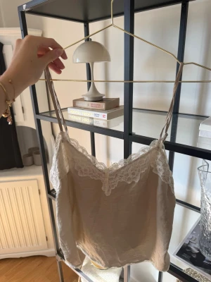 Beige linnetopp med spets från Stradivarius - Säljer en beige linnetopp från Stradivarius med tunna axelband och vacker spetsdetalj längs urringningen. Toppen har en mjuk, silkeslen känsla och är perfekt för att skapa en chic och avslappnad look.🤍