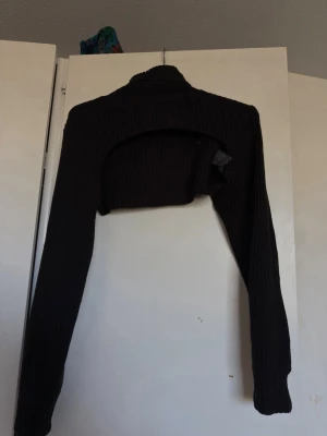 Svart stickad bolero - Säljer en svart stickad bolero med lång ärm och ribbad struktur. Perfekt att slänga över en topp för extra värme och stil. Kort modell som slutar ovanför midjan och har en tajt passform.
