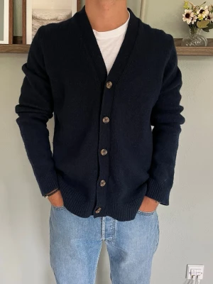 Dobber Cardigan Mörkblå M - Dobber cardigan i mörkblått med stilren och klassisk design. Stickad cardigan med V-ringning, träfärgade knappar och ribbade muddar. Perfekt över en t-shirt eller skjorta och passar enkelt med jeans eller chinos.  Skick: fint skick, inga hål eller fläckar Storlek: M Modell: 175 cm / 65 kg — sitter true to size på bild