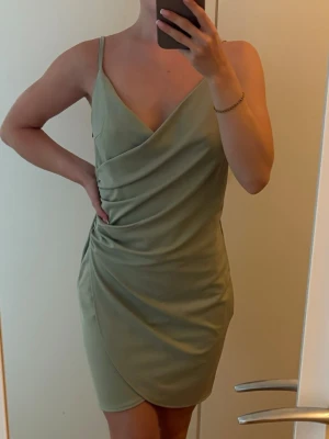 Ljusgrön omlottklänning med smala band - Snygg ljusgrön kort omlottklänning med smala axelband och v-ringning. Klänningen har draperade detaljer framtill och en figurnära passform som ger en elegant siluett. Oanvänd men tagit bort prislappen. 