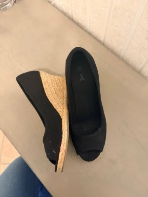 Svarta kilklackade espadrille-pumps - Säljer ett par svarta pumps med öppen tå och kilklack i flätad jute. Skorna har en klassisk espadrille-look och är perfekta för dig som vill ha både stil och komfort. Materialet är tyg och sulan har en snygg naturfärgad detalj.