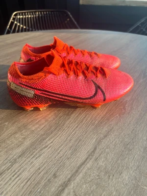 Nike mercurial Vapor 13 elite SG  - Säljer ett par röda Nike Mercurial fotbollsskor i stl 37,5 med svart swoosh och detaljer i guld. Skorna har låg profil, snörning och ovandel i syntetmaterial som ger bra bollkontroll. Perfekta för dig som vill sticka ut på planen!